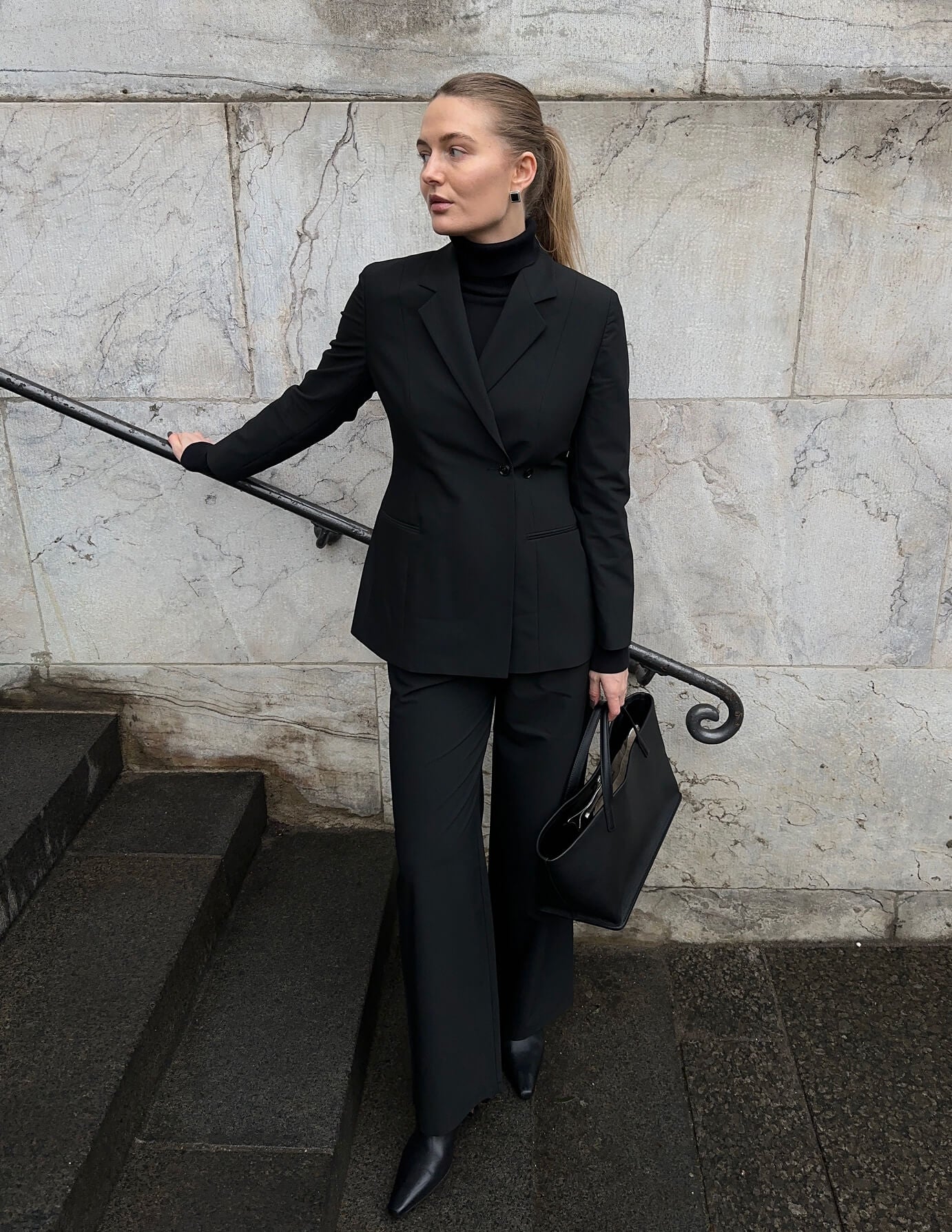 A woman wearing a Women’s Avira Blazer Black and matching trousers stands on stone steps holding a black tote bag, embodying a minimalist Scandinavian design aesthetic.